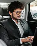 Un hombre con traje y gafas sentado en el asiento trasero de un coche, usando una laptop.